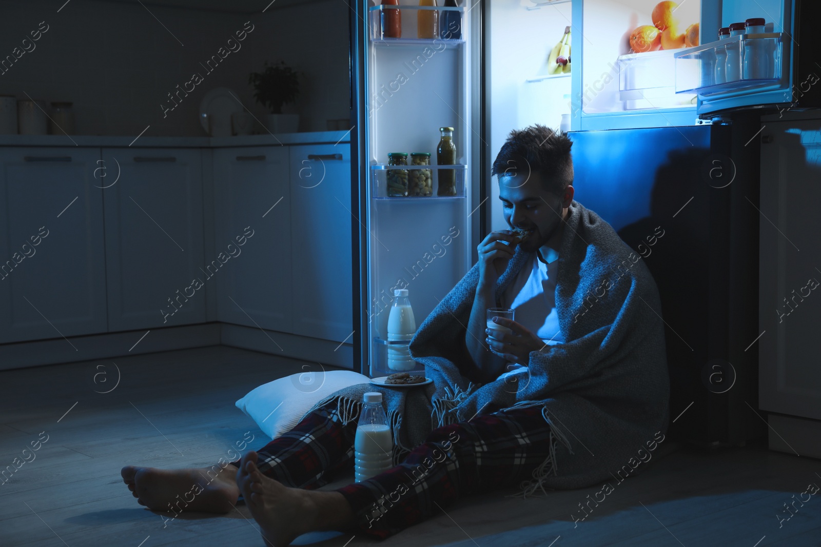 Photo of Young man eating cookies near open refrigerator in kitchen at night