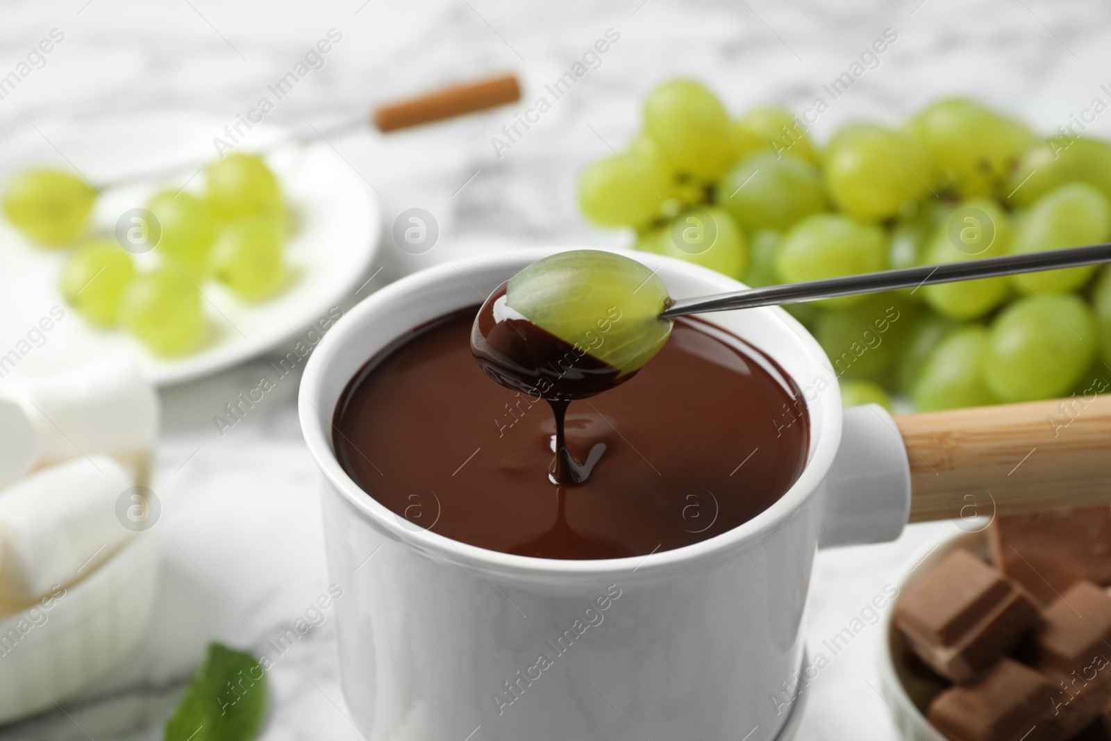 Photo of Dipping grape into fondue pot with dark chocolate on marble table