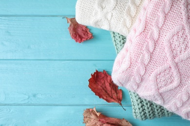 Stack of folded warm sweaters and dry leaves on turquoise wooden background, top view. Space for text