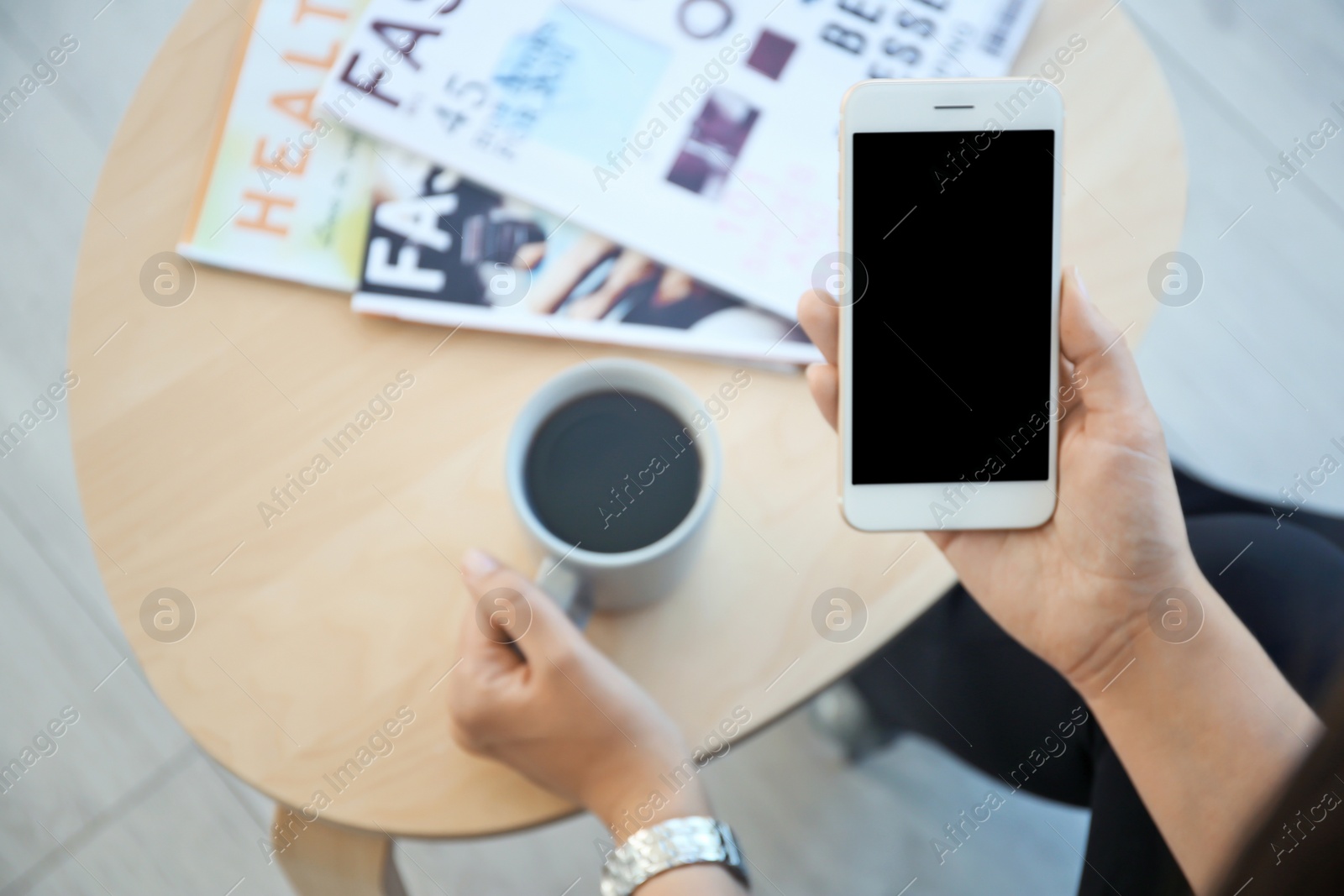 Photo of Young woman with cup of coffee holding mobile phone in hand at table indoors