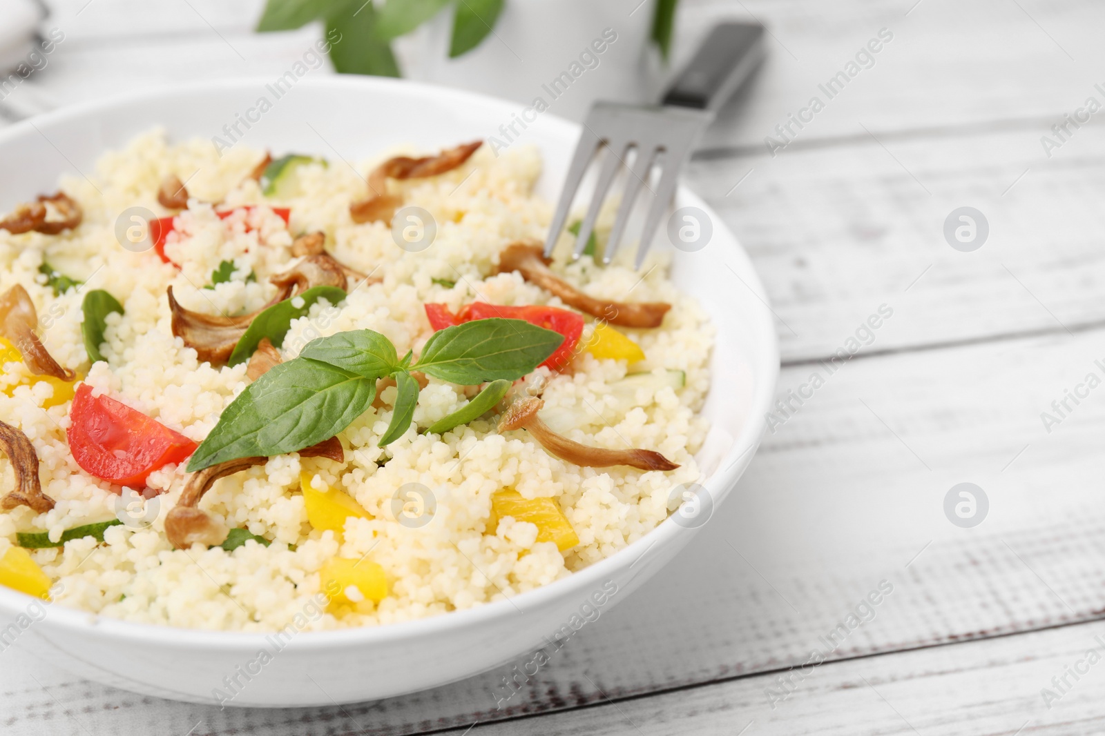 Photo of Bowl of delicious couscous with vegetables and basil served on white wooden table, closeup. Space for text