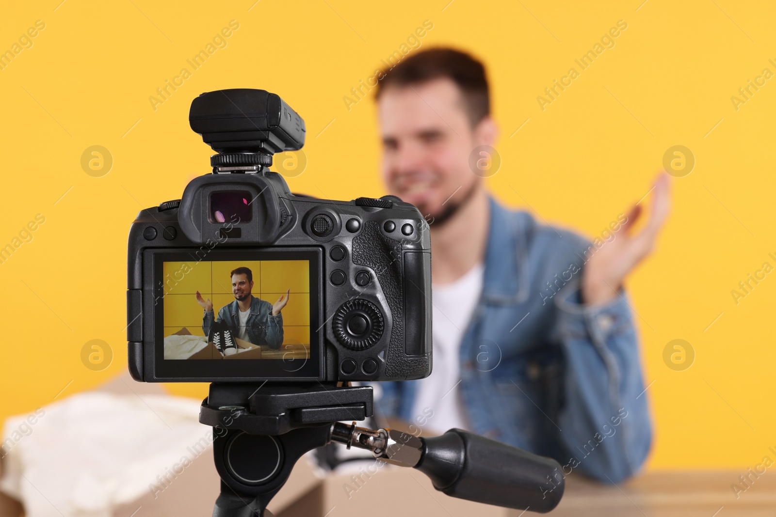 Photo of Fashion blogger showing sneakers while recording video at table against orange background, focus on camera