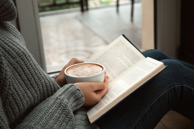 Photo of Woman with cup of coffee reading book near window indoors, closeup