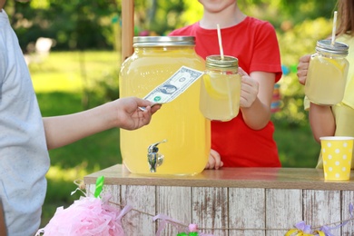 Photo of Little girls selling natural lemonade to boy in park, closeup. Summer refreshing drink
