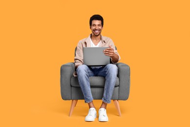 Happy man with laptop sitting in armchair on orange background