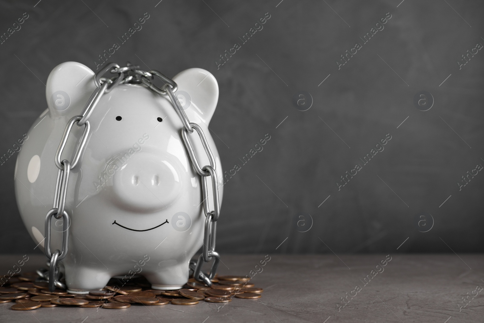 Photo of Piggy bank with steel chain and coins on grey stone table, space for text. Money safety concept