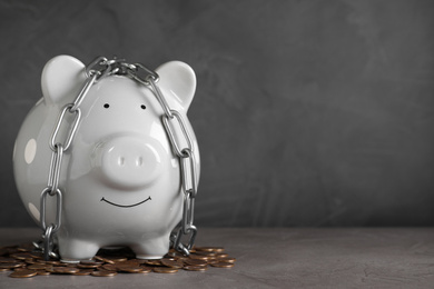 Photo of Piggy bank with steel chain and coins on grey stone table, space for text. Money safety concept