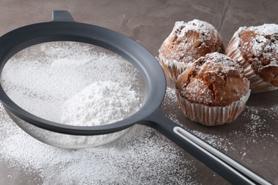 Sieve with sugar powder and muffins on grey table, closeup