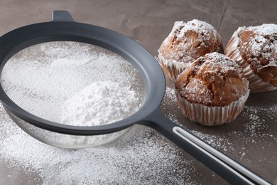 Photo of Sieve with sugar powder and muffins on grey table, closeup