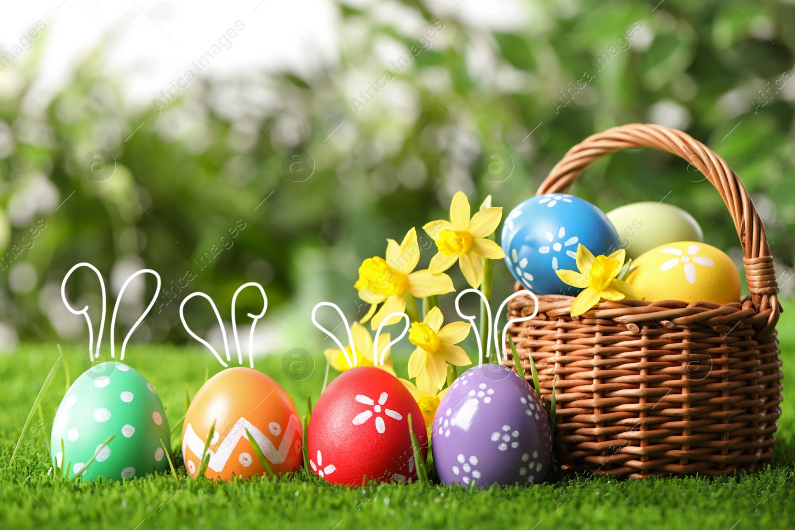 Image of Easter eggs with cute bunny ears, near wicker basket and flowers on green grass