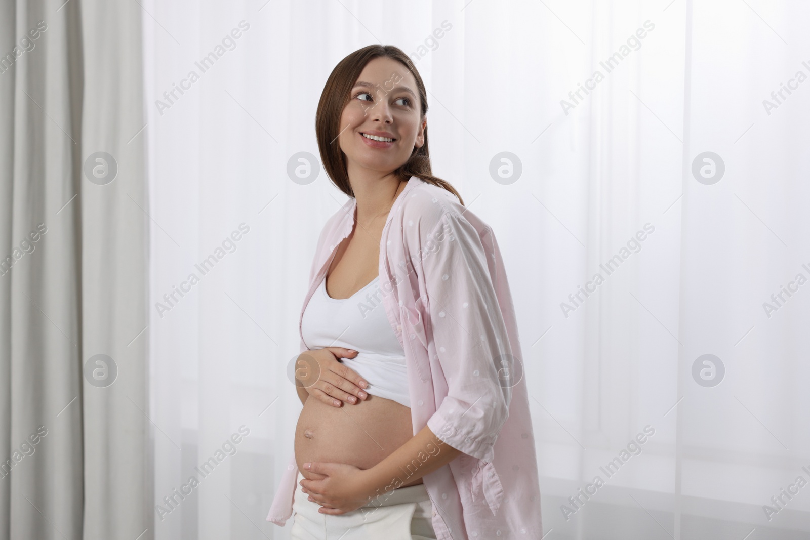 Photo of Beautiful pregnant woman in pink shirt near window indoors