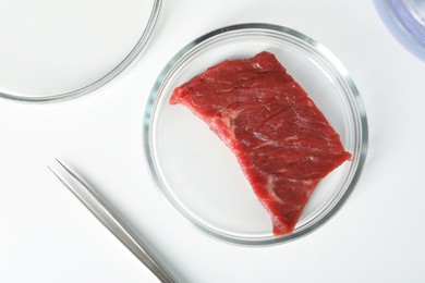 Petri dish with piece of raw cultured meat and tweezers on white table, flat lay