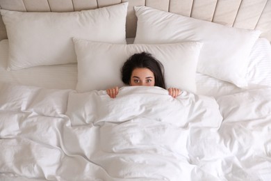 Young woman hiding under warm white blanket in bed, top view