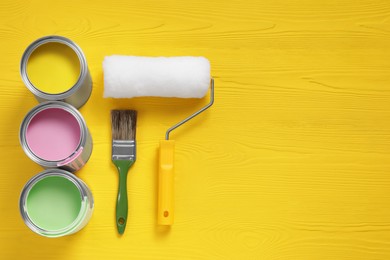 Photo of Cans of paints, brush and roller on yellow wooden table, flat lay. Space for text