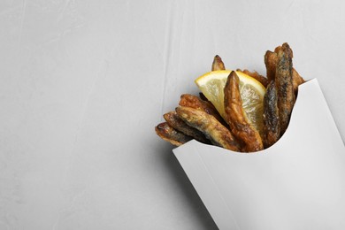 Photo of Paper bag with delicious fried anchovies and lemon slice on light table, top view. Space for text