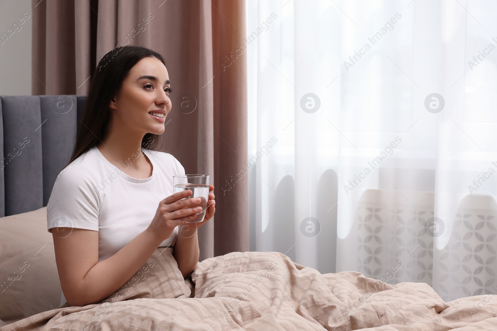 Photo of Beautiful woman with glass of water on bed at home, space for text. Lazy morning