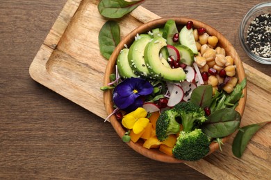 Delicious vegan bowl with broccoli, avocados and violet flowers on wooden table, flat lay