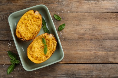 Photo of Halves of cooked spaghetti squash in baking dish, basil and salt on wooden table, flat lay. Space for text