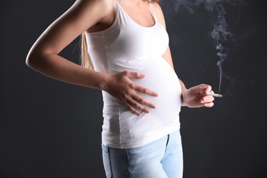 Photo of Young pregnant woman smoking cigarette on dark background, closeup. Harm to unborn baby