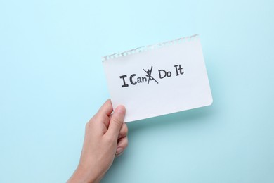 Photo of Motivation concept. Woman holding paper with changed phrase from I Can't Do It into I Can Do It by crossing over letter T on light blue background, top view