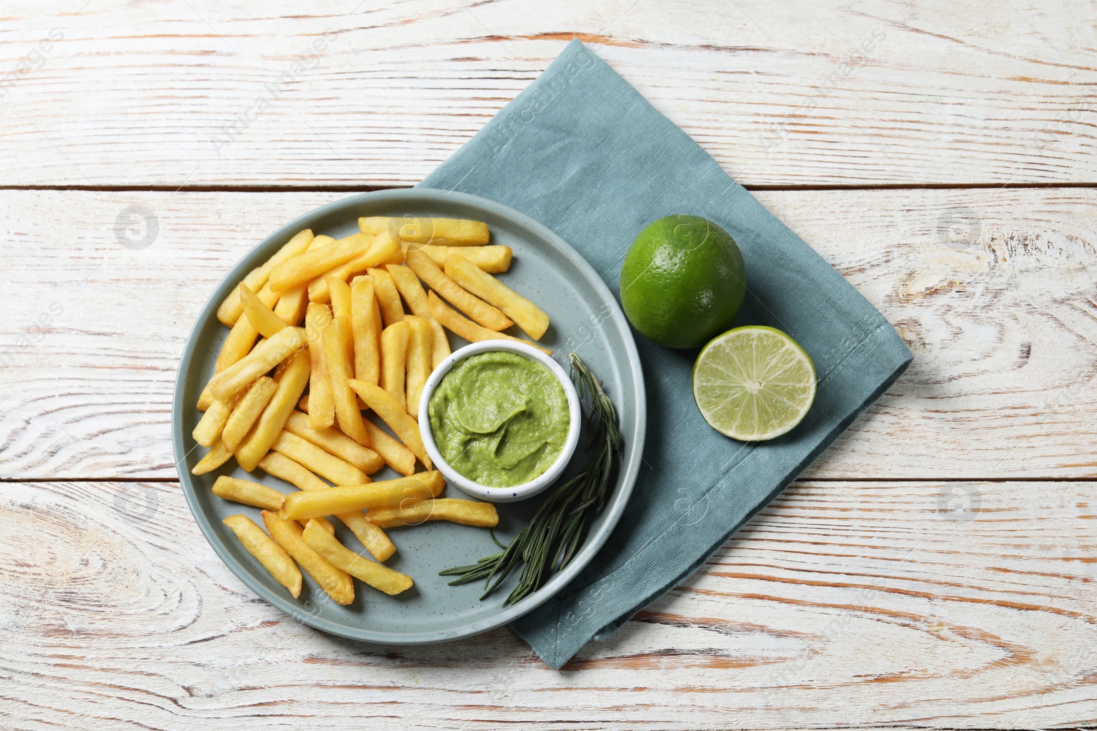 Photo of Plate with delicious french fries, avocado dip, lime and rosemary served on white wooden table, top view