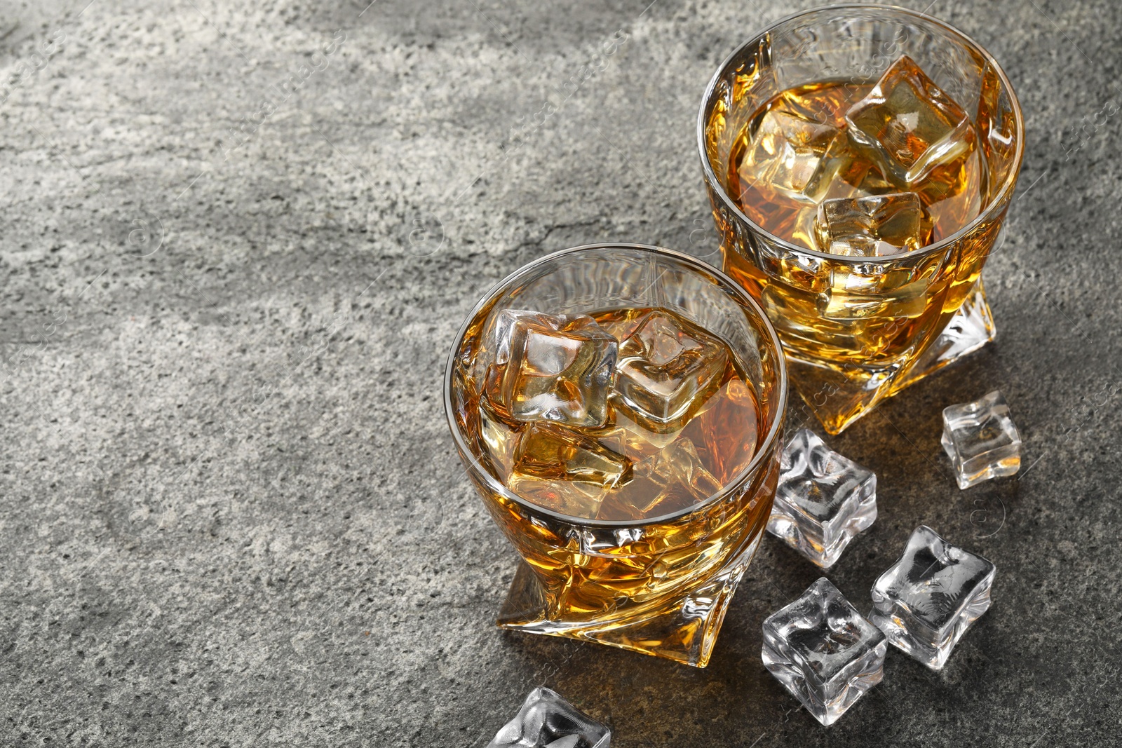 Photo of Whiskey and ice cubes in glasses on grey textured table, space for text