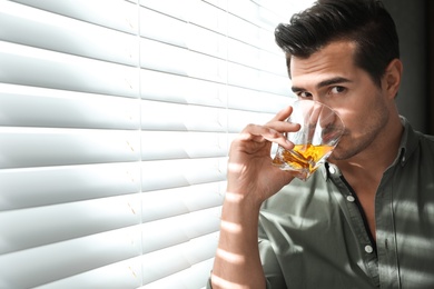 Young man with glass of whiskey near window indoors. Space for text