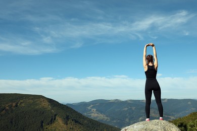 Beautiful young woman stretching on rock in mountains, back view