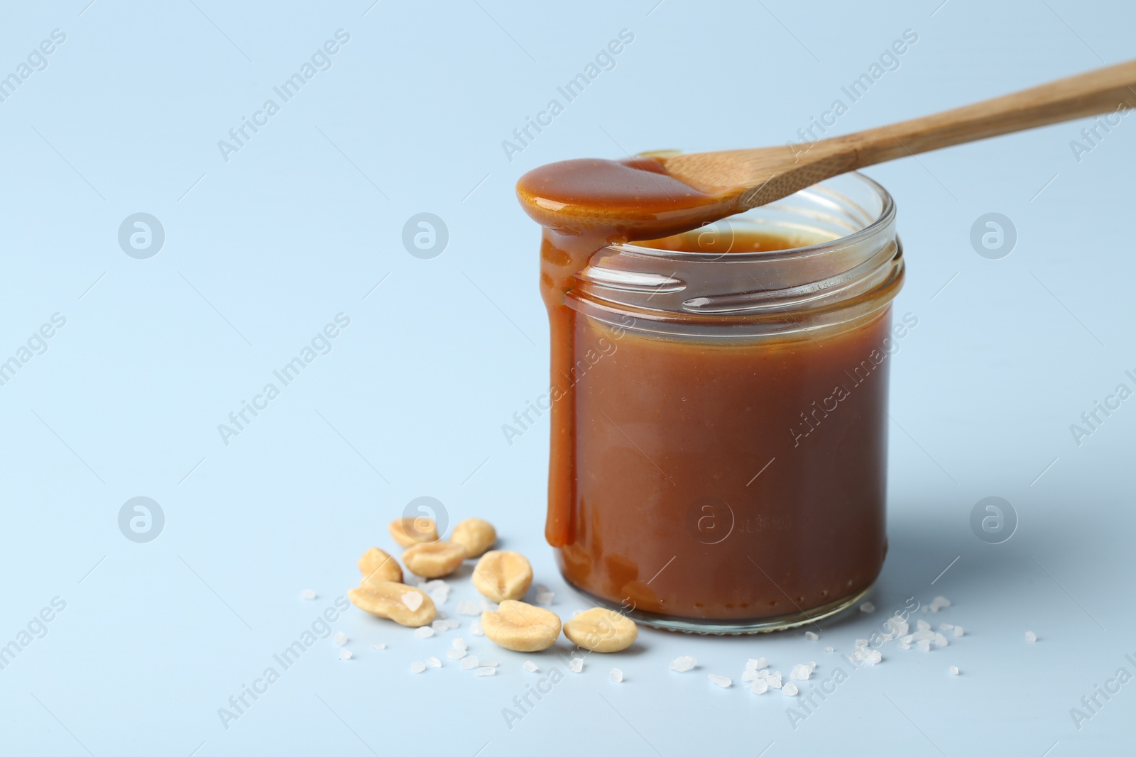 Photo of Jar of tasty salted caramel, spoon and peanuts on light blue background, closeup. Space for text