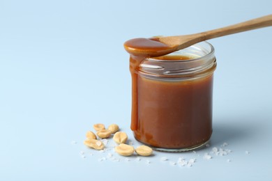 Photo of Jar of tasty salted caramel, spoon and peanuts on light blue background, closeup. Space for text