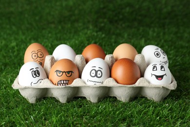Photo of Eggs with different drawn faces in cardboard package on green grass