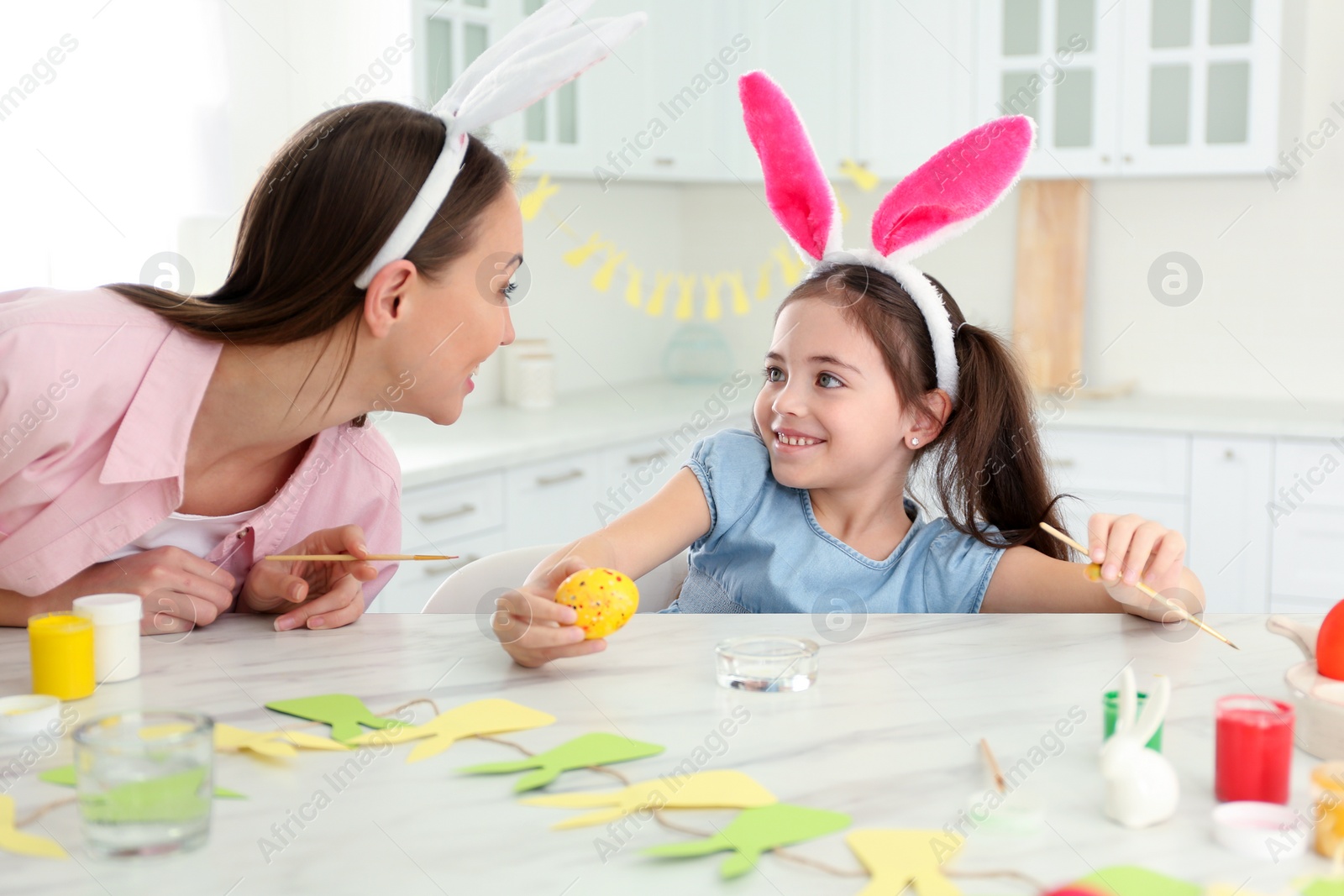 Photo of Mother and daughter with bunny ears headbands painting Easter egg in kitchen