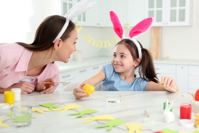 Photo of Mother and daughter with bunny ears headbands painting Easter egg in kitchen