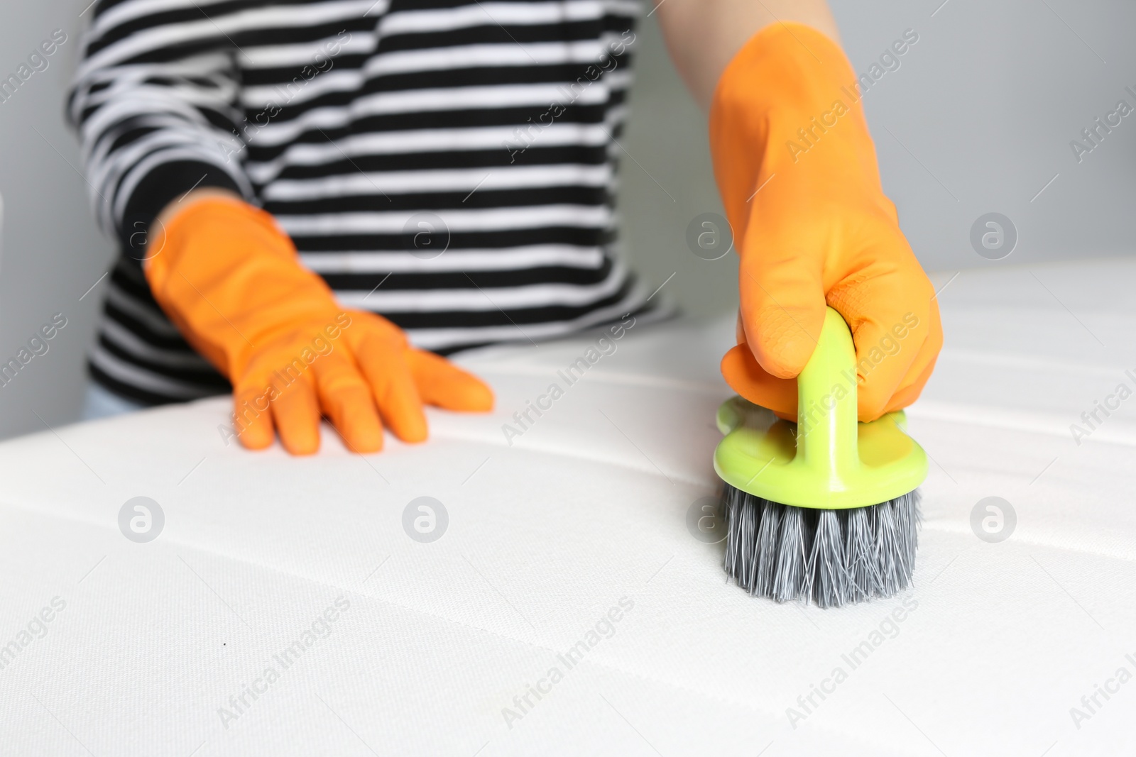 Photo of Woman in orange gloves cleaning white mattress with brush indoors, closeup