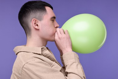 Photo of Young man inflating light green balloon on purple background