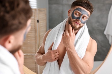 Photo of Handsome man with clay mask on his face near mirror in bathroom