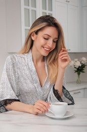 Photo of Pretty young woman in beautiful silk robe with cup of coffee at kitchen table