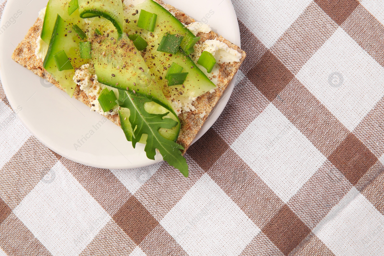 Photo of Fresh crunchy crispbread with cream cheese, cucumber, green onion and arugula on checkered tablecloth, top view. Space for text