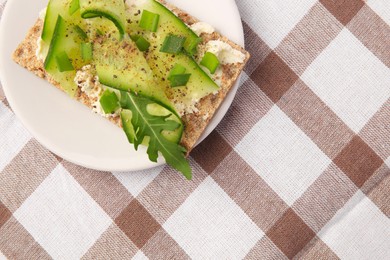 Fresh crunchy crispbread with cream cheese, cucumber, green onion and arugula on checkered tablecloth, top view. Space for text