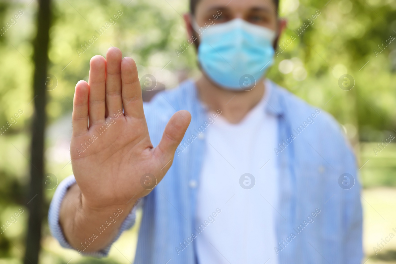 Photo of Man in protective face mask showing stop gesture in park, closeup. Prevent spreading of coronavirus