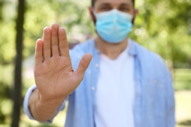 Photo of Man in protective face mask showing stop gesture in park, closeup. Prevent spreading of coronavirus