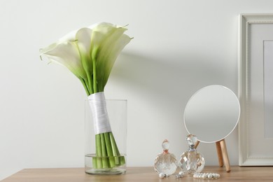 Beautiful calla lily flowers in glass vase, bottles with perfumes, jewelry and mirror on wooden table