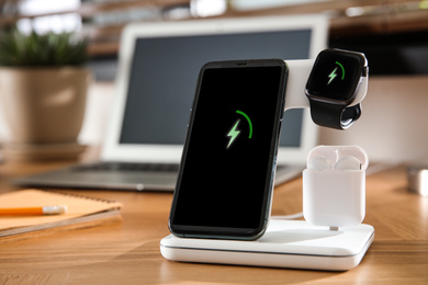 Photo of Mobile phone, earphones and smartwatch charging with wireless pad on wooden desk. Modern workplace device