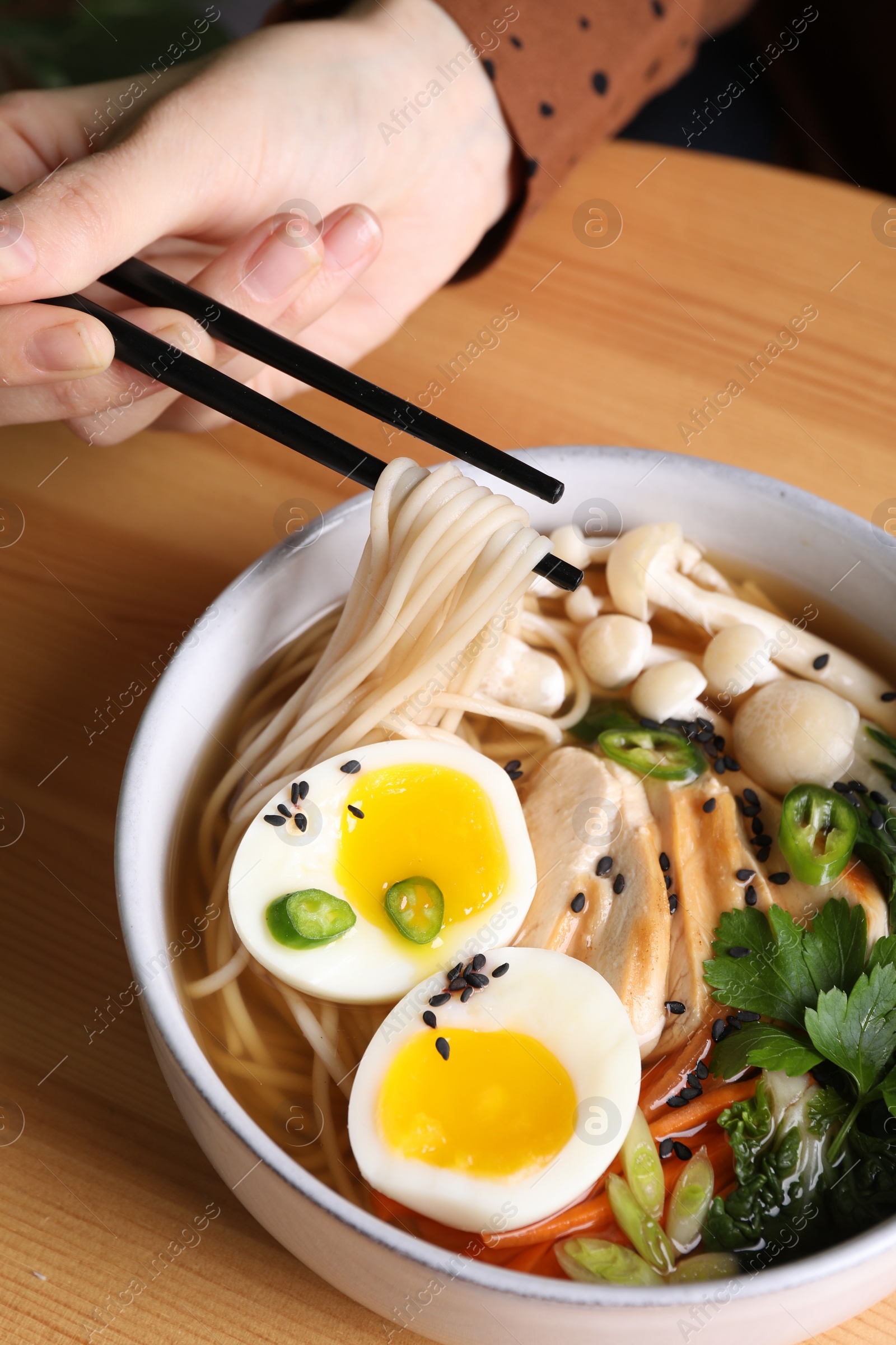 Photo of Woman eating delicious ramen with chopsticks at wooden table, closeup. Noodle soup