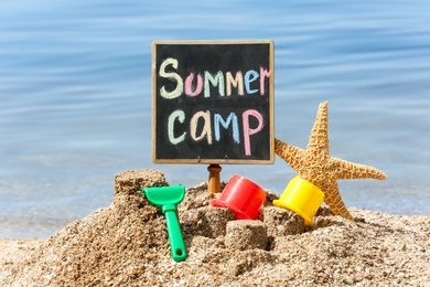 Photo of Little blackboard with text SUMMER CAMP and toys on sand near sea