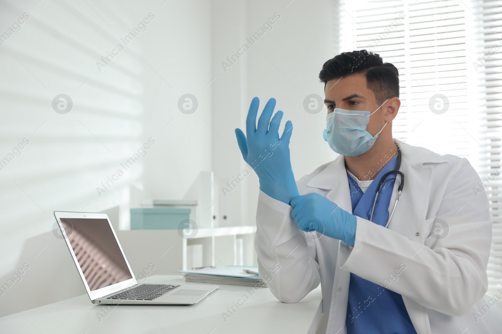 Photo of Doctor in protective mask putting on medical gloves at table in office