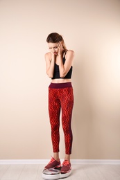 Sad young woman measuring her weight using scales on floor near light wall. Weight loss motivation