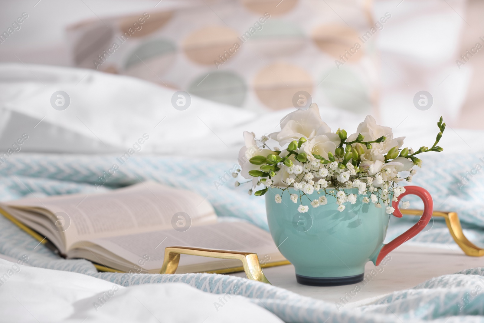 Photo of Beautiful bright flowers in cup, open book and tray on bed. Space for text