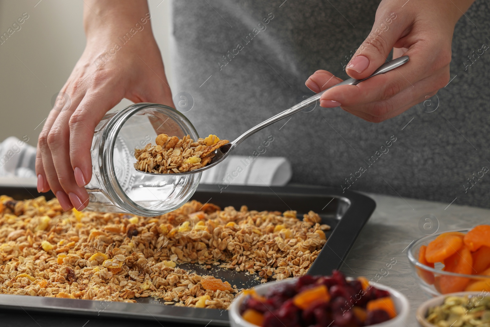 Photo of Woman putting granola from baking tray into jar at grey table, closeup