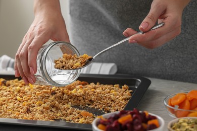 Woman putting granola from baking tray into jar at grey table, closeup